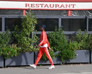 Pierre · 18/10/2025 · 3469 x 2808 Cette photographie montre une personne vêtue d’une combinaison rouge, marchant devant un restaurant. Le sujet principal est cette silhouette en mouvement, légèrement floue, suggérant dynamisme.La composition est simple, avec un cadrage serré sur la personne et le bâtiment en arrière-plan. Les couleurs dominantes sont le rouge vif de la combinaison et le vert des plantes grimpantes. La lumière semble naturelle, probablement en journée, avec des ombres douces.L’ambiance est urbaine et moderne, avec une touche de minimalisme et de contraste entre les couleurs vives et les tons neutres du bâtiment.
