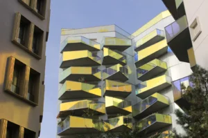 Marie-Odile · 1600 x 1066 Cette photographie met en scène un immeuble moderne aux façades géométriques et colorées. Le sujet principal est ce bâtiment aux formes anguleuses, recouvertes de panneaux jaunes et transparents, créant un effet dynamique.La composition joue sur les contrastes entre les lignes droites et les angles variés. Le cadrage, légèrement bas, accentue la hauteur et la verticalité de l'édifice. La lumière, naturelle et vive, souligne les reflets et les transparences des panneaux.Les couleurs dominantes sont le jaune vif et les teintes bleutées des vitres, apportant une touche futuriste. Le style architectural est contemporain et audacieux, évoquant une ambiance innovante et urbaine.
