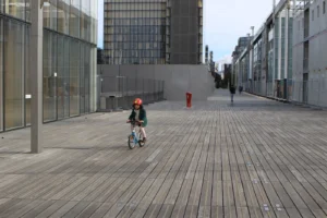 Marie-Odile · 1600 x 1066 Cette photographie montre un enfant en combinaison rouge et jaune roulant un petit vélo sur une allée en bois.La composition est épurée, avec un cadrage serré sur le sujet, mettant en valeur l’espace urbain moderne autour. Les bâtiments en verre et béton dominent l’arrière-plan. La lumière, naturelle et diffuse, crée des ombres douces. Les couleurs dominantes sont les tons neutres (gris, beige) avec des touches vives (rouge, jaune) apportées par la combinaison de l’enfant.L’ambiance est à la fois calme et dynamique, mêlant innocence et modernité architecturale.