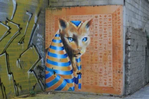 Marie-Odile · 1600 x 1066 Cette photographie met en scène un **renard stylisé en tête d'animal égyptien**, probablement un chacal, avec des **bandes bleues et orange** et des yeux bleus perçants. La **composition** est centrée sur ce motif, encadré par un **art mural coloré** aux motifs géométriques et symboles hiéroglyphiques.Le **cadrage** est serré, mettant en valeur les détails. La **lumière** semble naturelle, éclairant le sujet de face. Les **couleurs dominantes** sont vives : orange, bleu, jaune et noir. Le **style** mêle **street art** et symbolisme égyptien, créant une **ambiance mystérieuse et artistique**, entre tradition et modernité.