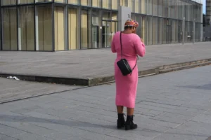 Marie-Odile · 1600 x 1066 Cette photographie met en scène une femme vêtue d’une robe rose fluo, sujet principal. Elle est cadrée de dos, en position légèrement penchée, tenant un sac noir et un parapluie.La composition est épurée, avec un premier plan dominé par la personne et un arrière-plan urbain neutre (bâtiments modernes, trottoir). La lumière semble naturelle, mais diffuse, sans ombres marquées. Les couleurs dominantes sont le rose vif de la robe, contrastant avec les tons gris et beiges de l’environnement.Le style est documentaire, avec une ambiance à la fois ordinaire et légèrement décalée, soulignée par le choix vestimentaire audacieux.