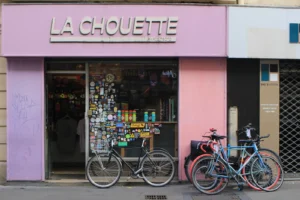 Marie-Odile · 1600 x 1066 Cette photographie montre une boutique nommée **« La Chouette »**, spécialisée en accessoires pour cyclistes. Le sujet principal est l’entrée colorée de la boutique, avec ses vélos exposés devant.La composition est équilibrée, avec un cadrage serré sur la façade. La lumière est naturelle et douce, mettant en valeur les couleurs vives. Les teintes dominantes sont le rose, le bleu et le violet, créant une ambiance joyeuse et moderne. Le style est urbain et décontracté, évoquant un commerce convivial et créatif.