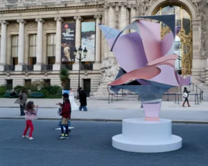 Cyrille · 24/10/2025 · 1500 x 1200 Cette photographie met en scène une grande sculpture abstraite et colorée, aux formes organiques et dynamiques, en tons de bleu, rose et blanc. Le sujet principal est cette œuvre, posée sur un socle circulaire.La composition associe cette sculpture à un cadre architectural imposant en arrière-plan, avec des colonnes et une façade classique. Le cadrage inclut aussi des passants, dont deux enfants en premier plan, ajoutant une touche de vie quotidienne.La lumière est naturelle, claire, et met en valeur les couleurs vives de la sculpture. L’ambiance est à la fois artistique et urbaine, mêlant modernité et tradition.