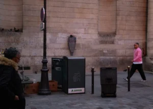 Muriel · 18/10/2025 · 2048 x 1463 Cette photographie montre une scène urbaine avec pour sujet principal un groupe d’objets et des passants en arrière-plan. La composition inclut des poubelles modernes et un lampadaire, avec une personne à gauche et une autre marchant à droite.Le cadrage est serré sur les éléments proches, créant une ambiance de rue quotidienne. La lumière est naturelle, légèrement tamisée, suggérant un jour couvert. Les couleurs dominantes sont les tons neutres (gris, beige) avec des touches de rouge et de vert. Le style est documentaire et réaliste, évoquant une ambiance calme et ordinaire dans un environnement urbain.