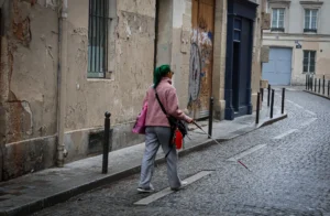 Muriel · 25/10/2025 · 2048 x 1338 Cette photographie montre deux personnes marchant dans une rue étroite et pavée, l’une portant un bonnet et un manteau rose, l’autre un sac rouge. Le sujet principal est cette femme en premier plan, dont le visage est flouté.La composition est simple, avec un cadrage serré sur les personnages et un arrière-plan de bâtiments vieillissants. La lumière est naturelle, tamisée, créant des ombres douces. Les couleurs dominantes sont les tons gris-beige des murs, rehaussés par les touches de rose et rouge des vêtements.Le style est réaliste et documentaire, évoquant une ambiance urbaine calme et légèrement mélancolique.
