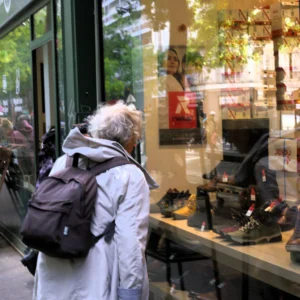 Fabienne · 3145 x 3145 Cette photographie montre une personne vue de dos, portant une veste blanche et un sac à dos, observant une vitrine de magasin. La composition met en valeur des chaussures exposées derrière la vitrine, reflétant une ambiance urbaine.Le cadrage est serré sur la personne et la vitrine, avec une légère flou artistique. La lumière est naturelle et douce, créant des contrastes subtils. Les couleurs dominantes sont les tons neutres (blanc, gris) et quelques éclats de couleurs vives des chaussures et des accessoires.L’ambiance est calme et quotidienne, typique d’une rue commerçante animée.