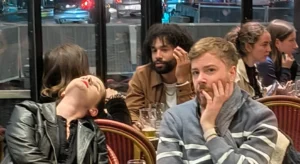 Laurence · 04/10/2025 · 1678 x 918 Cette photographie capture un groupe de personnes assises dans un lieu public, probablement un café ou un restaurant. Le sujet principal semble être les trois individus au premier plan, dont deux hommes et une femme, tous avec des expressions de fatigue ou d’ennui.La composition est serrée, avec un cadrage rapproché qui met l’accent sur les visages. La lumière est douce et naturelle, sans ombres marquées, suggérant un éclairage intérieur standard. Les couleurs dominantes sont neutres et sobres, avec des tons gris, beiges et quelques touches de couleurs vives portées par les vêtements.L’ambiance est détendue mais un peu morose, reflétant une scène de vie quotidienne.