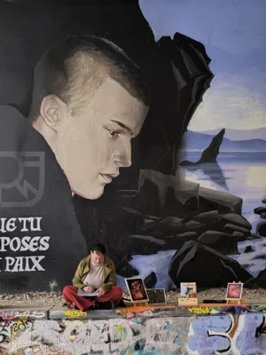 Laurence · 04/10/2025 · 3060 x 4080 Cette photographie montre un mural représentant un visage masculin en gros plan, partiellement détruit, avec des éléments de graffiti et de tags autour. Au premier plan, une table est couverte de magazines, de livres et d’objets divers, évoquant un espace de travail ou de création artistique.Le cadrage est serré sur le visage et la table, créant un contraste entre destruction et création. Les couleurs dominantes sont des tons froids (bleus, gris) et des touches chaudes (rouges, jaunes) sur les objets. La lumière semble naturelle, avec des ombres douces. L’ambiance est à la fois mélancolique et dynamique, reflétant un mélange de désordre et de créativité.