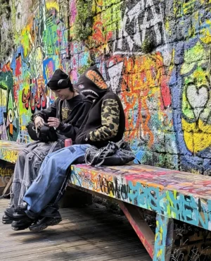 Laurence · 04/10/2025 · 2731 x 3371 Cette photographie montre un homme assis seul sur un banc coloré, absorbé par son téléphone. Le sujet principal est mis en valeur par un cadrage serré et une composition centrée.L'arrière-plan est un mur couvert de graffitis multicolores, apportant une ambiance urbaine et artistique. La lumière semble naturelle, douce et légèrement tamisée, créant des contrastes modérés. Les couleurs dominantes sont vives et variées grâce aux tags, tandis que les tons neutres du sujet (vêtements sombres) contrastent avec cet éclat.Le style est réaliste et spontané, évoquant une ambiance à la fois calme et dynamique.