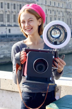Cédric · 11/10/2025 · 3059 x 4588 Cette photographie met en scène une jeune femme souriante, sujet principal, tenant une grosse caméra vintage à l'épaule. La composition est simple et centrée, avec un cadrage serré sur elle. L'arrière-plan montre un canal et des bâtiments, suggérant un cadre urbain.La lumière est naturelle et douce, éclairant son visage avec des ombres légères. Les couleurs dominantes sont les tons chauds (peau claire, rouge du bonnet) et les teintes neutres (gris de la caméra et du fond). Le style est documentaire et spontané, évoquant une ambiance joyeuse et créative.