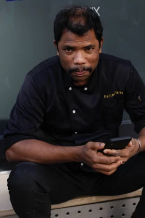 Cédric · 11/10/2025 · 3170 x 4755 Cette photographie montre un homme assis, vêtu d'une tenue noire professionnelle (polo et pantalon), en train de consulter son téléphone portable. Le cadrage est serré, centré sur son visage et ses mains. La lumière semble naturelle et douce, sans ombres marquées.Les couleurs dominantes sont les tons sombres (noir, gris) avec des touches neutres. L’ambiance est sobre et professionnelle, évoquant un environnement de travail ou une interview. Le style est réaliste et direct, sans mise en scène particulière.