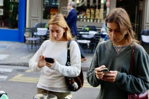 Cédric · 11/10/2025 · 5184 x 3456 Cette photographie montre deux jeunes femmes comme sujets principaux, toutes deux absorbées par leurs smartphones. La composition est dynamique, avec un cadrage serré sur elles en mouvement, en extérieur.La lumière est naturelle, probablement en journée, avec des ombres douces. Les couleurs dominantes sont neutres et douces : gris (vêtements, trottoir), beige et quelques touches de couleurs vives (sac, détails urbains).Le style est documentaire et spontané, capturant un moment du quotidien. L’ambiance est urbaine et moderne, reflétant une vie trépidante et connectée.