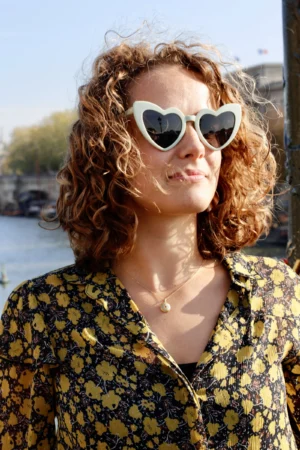 Cédric · 11/10/2025 · 3456 x 5184 Cette photographie met en scène une femme aux cheveux bouclés comme sujet principal. Elle porte des lunettes à forme de cœur et un chemisier à motifs floraux jaunes et verts.Le cadrage est serré, centré sur son visage et sa poitrine. La lumière naturelle, douce et légèrement latérale, crée des ombres douces et met en valeur les détails. Les couleurs dominantes sont les tons chauds (jaunes, verts) et les contrastes avec sa peau claire.Le style est décontracté et estival, évoquant une ambiance détendue et joyeuse, presque bohème.