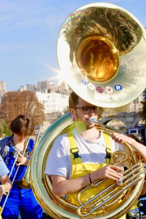 Cédric · 11/10/2025 · 3456 x 5184 Cette photographie met en scène un musicien jouant du tuba, instrument principal. La composition montre aussi partiellement une autre personne jouant du trombone.Le cadrage est serré sur le tuba et le musicien, avec un arrière-plan flou de bâtiments et de ciel. La lumière est naturelle et semble ensoleillée, créant des contrastes nets. Les couleurs dominantes sont les tons dorés du tuba, les verts et gris de l’uniforme, et les teintes bleutées du ciel.Le style est dynamique et spontané, évoquant une ambiance festive et artistique, probablement lors d’un concert en extérieur.