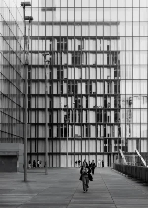 Philippe · 18/10/2025 · 2143 x 3000 Cette photographie met en scène deux personnes marchant devant un immeuble moderne. Le sujet principal est l’architecture, avec ses grandes fenêtres en miroir reflétant le ciel et les structures environnantes. La composition joue sur les lignes verticales et horizontales, créant une symétrie et une profondeur.Le cadrage est serré sur le bâtiment, avec une lumière naturelle douce, typique d’un jour couvert. Les couleurs dominantes sont les tons de gris, renforçant l’aspect urbain et minimaliste. Le style est épuré et contemporain, évoquant une ambiance calme et réfléchie, presque mélancolique.