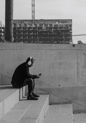Philippe · 18/10/2025 · 2094 x 3000 Cette photographie en noir et blanc met en scène un homme assis seul sur des marches, le regard dirigé vers le bas, absorbé par son téléphone portable. La composition est épurée, avec un cadrage serré sur le sujet, isolé devant un bâtiment industriel en arrière-plan.La lumière est douce, sans contraste marqué, créant une ambiance sobre et mélancolique. Les tons gris dominent, renforçant une atmosphère contemplative et introspective. Le style est documentaire et minimaliste, évoquant une solitude urbaine moderne.