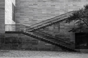 Philippe · 18/10/2025 · 3000 x 2001 Cette photographie en noir et blanc met en scène un **escalier extérieur en béton** bordé de rampes métalliques, sujet principal. La **composition** repose sur des lignes diagonales convergentes, créant une perspective dynamique. Le **cadrage** serré isole l’escalier, renforçant son importance.La **lumière** douce et naturelle, probablement matinale ou vesprinale, adoucit les contrastes. Les **couleurs dominantes** sont les nuances de gris, accentuées par l’absence de couleur.Le style est **minimaliste et géométrique**, évoquant une ambiance **sobre et contemplative**, presque mélancolique. L’ombre et la lumière soulignent la texture des matériaux.