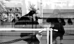 Pascale · 18/10/2025 · 2000 x 1196 Cette photographie en noir et blanc montre un sujet principal flou, probablement une personne en mouvement, reflétée dans une vitre. La composition joue sur les reflets et les superpositions, créant une ambiance mystérieuse et dynamique. Le cadrage est serré, mettant en valeur les détails des silhouettes et des éléments urbains.La lumière semble naturelle, mais diffuse, adoucissant les contrastes. Les tons dominants sont les gris et noirs, renforçant l’aspect urbain et moderne. Le style est épuré et artistique, évoquant une atmosphère à la fois mélancolique et vivante.