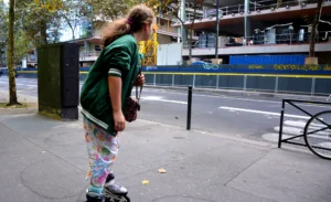 Pascale · 18/10/2025 · 2000 x 1222 Cette photographie montre une personne en train de faire du skate ou du roller dans un cadre urbain. Le sujet principal est une jeune fille vêtue d’un haut vert et de pantalons colorés, penchée en avant.La composition est dynamique, avec un cadrage serré sur le sujet. L’arrière-plan inclut des éléments urbains comme des barrières et des bâtiments. La lumière semble naturelle, légèrement tamisée, créant des ombres douces. Les couleurs dominantes sont les tons verts, gris et pastel des vêtements et de l’environnement.Le style est documentaire et spontané, évoquant une ambiance décontractée et moderne.