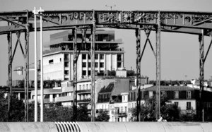 Pascale · 18/10/2025 · 2000 x 1249 Cette photographie en noir et blanc met en scène un **pont métallique** imposant en sujet principal, avec une structure industrielle et des poutres robustes. En arrière-plan, des **bâtiments urbains** partiellement construits ou en rénovation sont visibles.La **composition** joue sur les contrastes entre l’architecture moderne et l’infrastructure ancienne. Le **cadrage** est serré, centré sur le pont, avec une perspective légèrement basse. La **lumière** est naturelle, douce, sans ombres marquées, créant une ambiance neutre et intemporelle. Les **tons dominants** sont les gris et noirs, renforçant l’aspect industriel et minimaliste. Le style est **documentaire**, évoquant une ambiance **urbaine et mélancolique**.