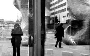 Pascale · 18/10/2025 · 2000 x 1250 Cette photographie en noir et blanc capture une scène urbaine vue à travers une vitre.Le **sujet principal** est une silhouette reflétée dans la vitre, partiellement visible, tandis qu’une personne réelle, vêtue d’un manteau, marche dehors. La **composition** joue sur le contraste entre intérieur et extérieur, avec un **cadrage** serré sur la vitre, créant une impression de profondeur.La **lumière** est naturelle et diffuse, adoucissant les contours. Les **couleurs dominantes** sont les nuances de gris, renforçant l’ambiance mélancolique et introspective.Le **style** est épuré et minimaliste, évoquant une **ambiance** contemplative et presque onirique.