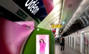 Pascale · 17/10/2025 · 2000 x 1226 Cette photographie montre une station de métro avec un train en mouvement à gauche. Le sujet principal est une affiche publicitaire lumineuse représentant une femme souriante. La composition est dynamique, avec un cadrage serré sur le train et l'affiche, et une perspective en profondeur vers le couloir de la station. La lumière est artificielle et uniforme, typique des stations de métro. Les couleurs dominantes sont le vert, le gris et le rose vif de l'affiche. Le style est urbain et moderne, créant une ambiance à la fois fonctionnelle et légèrement futuriste.