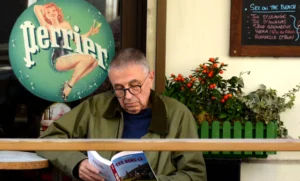 Pascale · 04/10/2025 · 2000 x 1207 Cette photographie montre un homme âgé lisant un livre, assis à une table. Le sujet principal est cet homme, dont le visage est partiellement caché par le livre.La composition inclut un fond avec une enseigne rétro "Perrier" et une plante fleurie. Le cadrage est serré sur le haut du corps de l’homme, avec une légère inclinaison vers le bas.La lumière semble naturelle et douce, créant une ambiance chaleureuse. Les couleurs dominantes sont des tons neutres (beige, vert) avec des touches de rouge et bleu sur l’enseigne. L’ambiance est calme et intime, évoquant un moment de détente.