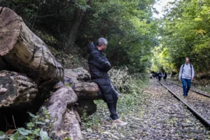 Benoit · 04/10/2025 · 5002 x 3335 Cette photographie montre un homme comme sujet principal, marchant sur une voie ferrée abandonnée, entouré de nature dense. La composition met en valeur un chemin sinueux bordé de rails et de végétation luxuriante. Le cadrage est serré sur l’homme au premier plan, avec une profondeur de champ qui inclut d’autres personnes en arrière-plan.La lumière semble naturelle et tamisée, typique d’un jour automnal. Les couleurs dominantes sont les verts profonds des feuilles et les tons terreux, avec des touches de brun et gris. Le style est documentaire et réaliste, créant une ambiance contemplative et mystérieuse, presque mélancolique.