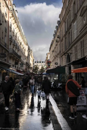 Benoit · 04/10/2025 · 5152 x 7728 Cette photographie capture une rue urbaine après une pluie, reflétant une ambiance humide et lumineuse. Le sujet principal est la rue bondée de passants, avec une perspective centrale en profondeur.La composition utilise un cadrage serré entre les bâtiments, créant un effet de tunnel. La lumière, douce et diffuse, provient probablement des nuages, adoucissant les contours. Les couleurs dominantes sont les tons gris-beige des bâtiments, contrastés par les reflets aquatiques et les éclats de rouge et bleu des enseignes et vêtements.Le style est réaliste et documentaire, évoquant une ambiance dynamique et vivante dans une ville européenne.