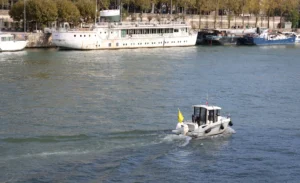 Laurence · 5881 x 3582 Cette photographie met en scène un petit bateau à moteur, sujet principal, navigant sur l’eau. Le cadrage est large, incluant aussi des bateaux ancrés en arrière-plan.La lumière semble naturelle, probablement en journée, avec des reflets sur l’eau. Les couleurs dominantes sont les bleus variés de l’eau et du ciel, contrastant avec les teintes grises et blanches des bateaux.Le style est documentaire, réaliste, sans effet artistique marqué. L’ambiance est calme et fonctionnelle, évoquant une activité portuaire ou de loisir.