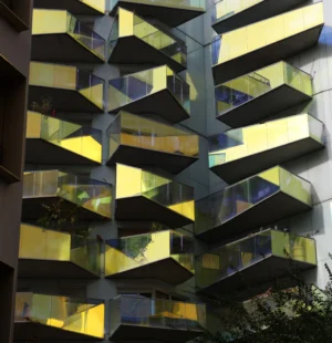 Laurence · 4485 x 4640 Cette photographie met en scène un immeuble moderne aux balcons en forme de losanges. Le sujet principal est l’architecture géométrique et les jeux de lumière sur les surfaces vitrées.La composition est symétrique et répétitive, avec des lignes horizontales et diagonales bien définies. Le cadrage est serré, centré sur la façade.Les couleurs dominantes sont des teintes froides (bleu, gris) et chaudes (jaune, doré) reflétées par les vitres. La lumière naturelle crée des reflets changeants, dynamiques.Le style est contemporain, minimaliste et architectural. L’ambiance est futuriste et presque abstraite, avec une touche de mouvement grâce aux reflets lumineux.