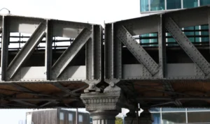 Laurence · 6960 x 4135 Cette photographie montre un **pont métallique** en structure complexe, sujet principal. La composition met en valeur les **poutres triangulées** et les supports en béton, soulignant l’ingénierie industrielle.Le cadrage serré isole la structure, avec une perspective légèrement basse. La lumière, naturelle et diffuse, adoucit les contours sans créer d’ombres marquées. Les **couleurs dominantes** sont les tons gris métallique et beige du béton, avec des touches de vert et de gris clair en arrière-plan.Le style est **documentaire et technique**, évoquant une ambiance urbaine et moderne, presque austère.
