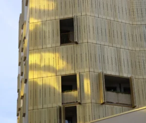 Laurence · 5494 x 4640 Cette photographie montre un immeuble moderne en façade de bois ou de matériau imitant le bois. Le sujet principal est la structure architecturale avec ses lignes verticales et ses fenêtres.Le cadrage est serré, mettant en valeur la texture et les jeux de lumière sur la surface. La lumière, naturelle et rasante, crée des ombres allongées et des reflets dorés, accentuant le relief.Les couleurs dominantes sont des tons bois clairs et des nuances dorées. Le style est épuré et contemporain, évoquant une ambiance industrielle et minimaliste, presque apaisante malgré l’aspect urbain.