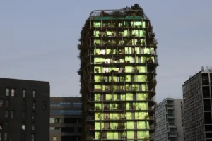 Laurence · 6960 x 4640 Cette photographie met en scène un **immeuble moderne vertical**, sujet principal, recouvert de végétation lumineuse. La **composition** est centrée, avec un cadrage serré qui isole le bâtiment parmi d'autres structures urbaines.La **lumière**, douce et naturelle, semble provenir des éclairages intérieurs, créant un contraste avec l'obscurité ambiante. Les **couleurs dominantes** sont les verts fluorescents et les teintes grises des autres bâtiments.Le style est **contemporain et architectural**, évoquant une ambiance **futuriste et écologique**, mêlant nature et urbanisme.