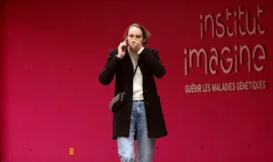 Franck T. · 10/10/2025 · 975 x 578 Cette photographie montre une femme comme sujet principal, vêtue d’un long manteau sombre et de jeans, discutant au téléphone devant un fond rouge. Le cadrage est serré, mettant en valeur son visage et sa silhouette.La lumière semble naturelle et douce, éclairant légèrement son visage. Les couleurs dominantes sont le rouge vif du fond et les tons neutres de ses vêtements. Le style est sobre et contemporain, évoquant une ambiance professionnelle ou scientifique, renforcée par le texte "Institut Imagine" en arrière-plan. L'atmosphère est calme et concentrée.