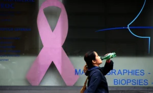 Franck T. · 10/10/2025 · 1000 x 607 Cette photographie met en scène un homme buvant une boisson, passant devant une vitrine. Le **sujet principal** est le contraste entre lui et l’**affiche médicale** derrière lui, montrant un ruban rose (symbole du cancer du sein) et des termes comme "mammographies" et "biopsies".La **composition** est simple, avec un **cadrage serré** sur l’homme et la vitrine. La **lumière** semble naturelle, bien que légèrement tamisée. Les **couleurs dominantes** sont le bleu (vêtement), le rose (ruban) et des teintes froides (arrière-plan médical). Le **style** est documentaire, évoquant une **ambiance urbaine et introspective**, mêlant vie quotidienne et enjeux de santé.