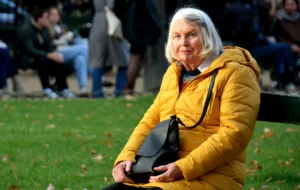 Pascale · 01/11/2025 · 1000 x 632 Cette photographie met en scène une femme âgée comme sujet principal, assise sur un banc dans un parc. Elle porte un manteau jaune vif et tient un sac à main.La composition est simple et naturelle, avec un cadrage serré sur elle, légèrement flou en arrière-plan. La lumière semble douce et naturelle, probablement en journée. Les couleurs dominantes sont les tons chauds (jaune, beige) contrastant avec les verts de la végétation.Le style est réaliste et spontané, évoquant une ambiance paisible et chaleureuse, presque intime.