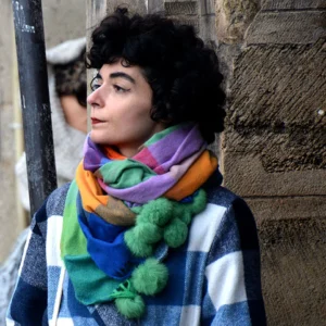 Pascale · 01/11/2025 · 1000 x 1000 Cette photographie met en scène une personne comme sujet principal, partiellement cadrée, avec un visage expressif et un regard pensif. Elle porte un pull à carreaux et un large écharpe multicolore aux couleurs vives (vert, orange, bleu, violet).La composition est simple, avec un cadrage serré sur le haut du corps. L'arrière-plan, flou et sombre, attire peu l'attention. La lumière semble naturelle, douce et légèrement tamisée, créant une ambiance chaleureuse et intime. Les couleurs dominantes sont vives et contrastées, apportant une touche joyeuse malgré le contexte sobre. Le style est réaliste et spontané.