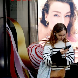 Pascale · 01/11/2025 · 1000 x 1000 Cette photographie montre une jeune femme comme sujet principal, absorbée par son téléphone portable. La composition joue sur le contraste entre elle et une grande affiche publicitaire derrière elle, représentant une autre femme aux yeux bleus.Le cadrage est serré, mettant en valeur la personne et son interaction avec la technologie. La lumière semble naturelle, bien que légèrement tamisée, créant des ombres douces. Les couleurs dominantes incluent des tons froids (gris, bleu) et des touches chaudes (rouge, blanc) sur l’affiche.Le style est urbain et contemporain, évoquant une ambiance moderne et quotidienne.