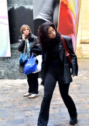 Pascale · 01/11/2025 · 703 x 1000 Cette photographie montre deux femmes en mouvement dans une rue pavée. La femme au premier plan, floue par le flou de mouvement, porte une veste noire et un sac bleu, marchant rapidement. L’arrière-plan révèle une autre femme, plus nette, tenant un téléphone et un sac à main.La composition met en valeur le dynamisme urbain. Le cadrage serré et l'angle bas accentuent le mouvement. La lumière naturelle semble douce mais contrastée, créant des ombres marquées. Les couleurs dominantes sont les tons neutres (noir, gris) avec des touches de bleu et rouge. Le style est documentaire et spontané, capturant une scène de vie quotidienne avec une ambiance vivante et moderne.