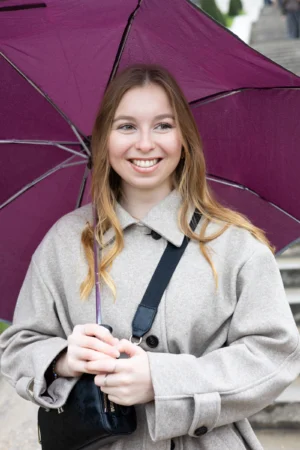 Claude · 4000 x 6000 Cette photographie met en scène une jeune femme souriante, sujet principal, tenant un parapluie violet. Le cadrage est serré, centré sur son visage et sa partie supérieure du corps. La lumière est naturelle et douce, éclairant son visage avec des ombres légères.Les couleurs dominantes sont les tons gris de son vêtement, contrastant avec le violet éclatant du parapluie. L’ambiance est joyeuse et détendue, renforcée par son sourire. Le style est simple et spontané, évoquant une scène de vie quotidienne.