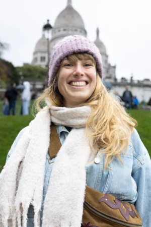 Claude · 3824 x 5736 Cette photographie met en scène une femme souriante comme sujet principal, vêtue d’un style décontracté et chaleureux. Elle porte un béret, un long foulard blanc et une veste légère, avec un sac en bandoulière.Le cadrage est serré sur son visage, avec en arrière-plan flou le Sacré-Cœur de Paris, suggérant un lieu emblématique. La lumière semble naturelle, douce et légèrement diffuse, créant une ambiance joyeuse. Les couleurs dominantes sont les tons pastel (bleu clair, gris) et le blanc du foulard, renforçant une impression de fraîcheur et de sérénité. L’ambiance est conviviale et touristique.