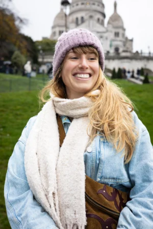 Claude · 4000 x 6000 Cette photographie met en scène une jeune femme souriante, sujet principal, vêtue d’un style décontracté et chaleureux. Elle porte un béret rose pâle, un manteau bleu clair, une écharpe et un sac en cuir.Le cadrage est serré sur elle, avec un arrière-plan flou montrant une architecture emblématique (le Sacré-Cœur de Paris) et un parc verdoyant. La lumière est naturelle et douce, probablement en journée.Les couleurs dominantes sont les tons pastel (rose, bleu) et les teintes automnales (beige, marron). L’ambiance est joyeuse et détendue, évoquant une balade agréable en ville.