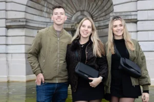 Claude · 5816 x 3877 Cette photographie montre trois jeunes posant ensemble devant une architecture en pierre.**Sujet principal** : Trois personnes souriantes, deux femmes et un homme, tenant des boîtes noires.
**Composition** : Alignement en triangle, cadrage serré sur les sujets.
**Lumière** : Naturelle et douce, éclairant uniformément les visages.
**Couleurs dominantes** : Tons neutres (beige, gris) avec des touches de vert (vêtements) et noir (boîtes).
**Style** : Informel et naturel, style documentaire.
**Ambiance** : Joyeuse et détendue, évoquant un moment de célébration ou de réussite.