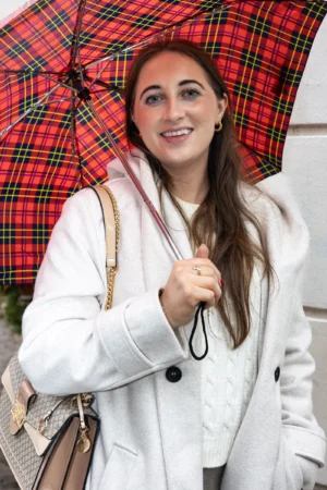 Claude · 3346 x 5019 Cette photographie met en scène une jeune femme souriante, sujet principal, tenant un parapluie à carreaux rouges et noirs. Elle porte un long manteau blanc et une écharpe beige, avec un sac en bandoulière.Le cadrage est serré, centré sur son visage et le haut de son corps. La lumière semble naturelle et douce, éclairant son visage avec une ambiance joyeuse. Les couleurs dominantes sont les tons blancs, rouges et noirs, avec des touches de beige.Le style est décontracté et élégant, évoquant une ambiance printanière ou automnale, chaleureuse et accueillante.