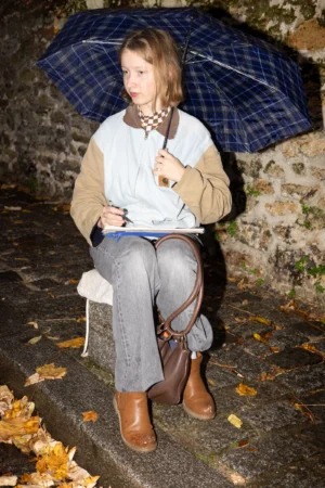 Claude · 4000 x 6000 Cette photographie met en scène une jeune femme comme sujet principal, assise sur un muret. Elle tient un parapluie bleu et plaqué, un carnet et un stylo, vêtue d’un style décontracté et élégant (pull, ceinture, jeans et bottines).La composition est simple et centrée, avec un cadrage serré sur elle. La lumière semble douce, probablement artificielle ou tamisée, créant une ambiance calme et intime. Les couleurs dominantes sont les tons neutres (beige, gris) rehaussés par les touches de bleu du parapluie et les reflets dorés des feuilles mortes. L’ambiance est à la fois studieuse et poétique, évoquant la réflexion ou la créativité.