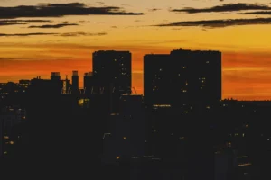Cette photographie capture un coucher de soleil en milieu urbain. Le **sujet principal** est la silhouette des **grands bâtiments** se découpant sur un ciel enflammé. La **composition** joue sur les contrastes entre les ombres noires des immeubles et les teintes chaudes du ciel.Le **cadrage** est large, offrant une vue panoramique de la ville. La **lumière**, douce et dorée, crée une ambiance apaisante et dramatique. Les **couleurs dominantes** sont les **oranges, rouges et jaunes** du ciel, opposés aux **noirs profonds** des bâtiments.Le style est **réaliste et contemplatif**, évoquant une **ambiance sereine et presque mélancolique**.