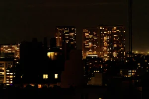Cette photographie capture un **paysage urbain nocturne**. Le **sujet principal** est une silhouette de gratte-ciels éclairés, formant une skyline. La **composition** oppose les bâtiments lumineux à l’obscurité ambiante, avec un arbre en premier plan.Le **cadrage** est large, incluant une vue d’ensemble de la ville. La **lumière**, douce et tamisée, met en valeur les éclairages des fenêtres et des enseignes. Les **couleurs dominantes** sont les tons **jaunes, oranges et noirs**, créant une ambiance **sombre et mystérieuse**.Le style est **réaliste et contemplatif**, évoquant une **ville endormie**, presque onirique.