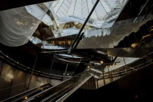 Cette photographie montre un escalier mécanique dans un espace intérieur moderne. Le sujet principal est l'escalier lui-même, avec ses structures métalliques et ses parois translucides.La composition est dynamique, avec un cadrage en contre-plongée accentuant la perspective. La lumière est douce mais contrastée, mettant en valeur les détails architecturaux. Les couleurs dominantes sont les tons froids (gris, blanc, bleu) et quelques éclats lumineux.Le style est épuré et contemporain, créant une ambiance futuriste et élégante, presque clinquante. L'atmosphère évoque un lieu public sophistiqué, comme un centre commercial ou un aéroport.