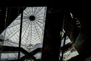 Cette photographie met en scène une **verrière géométrique** comme sujet principal, avec ses structures métalliques et ses vitraux formant des motifs rayonnants. La **composition** joue sur les lignes convergentes et la symétrie, créant une perspective profonde.Le **cadrage** est serré, accentuant les détails architecturaux. La **lumière**, tamisée et filtrée par les vitraux, génère des contrastes forts entre ombres et éclats lumineux. Les **couleurs dominantes** sont des teintes froides et neutres, avec des reflets bleutés et gris.Le style est **minimaliste et industriel**, évoquant une ambiance **mystérieuse et contemplative**, presque sacrée.